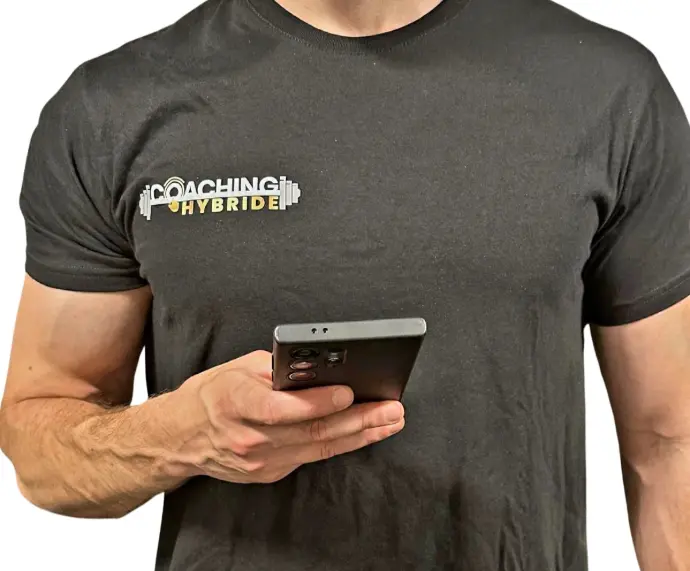 alt=&#34;Coach sportif tenant un smartphone avec l&#39;application Coaching Hybride, vêtu d&#39;un t-shirt à l&#39;effigie de sa marque&#34;